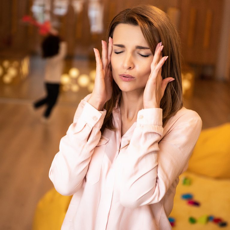 tired educator with closed eyes touching head in kindergarten, kid running in room
