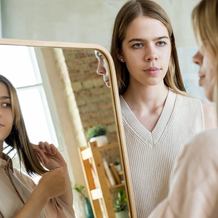 Serious teenage girl looking at her twin sister brushing hair in front of mirror